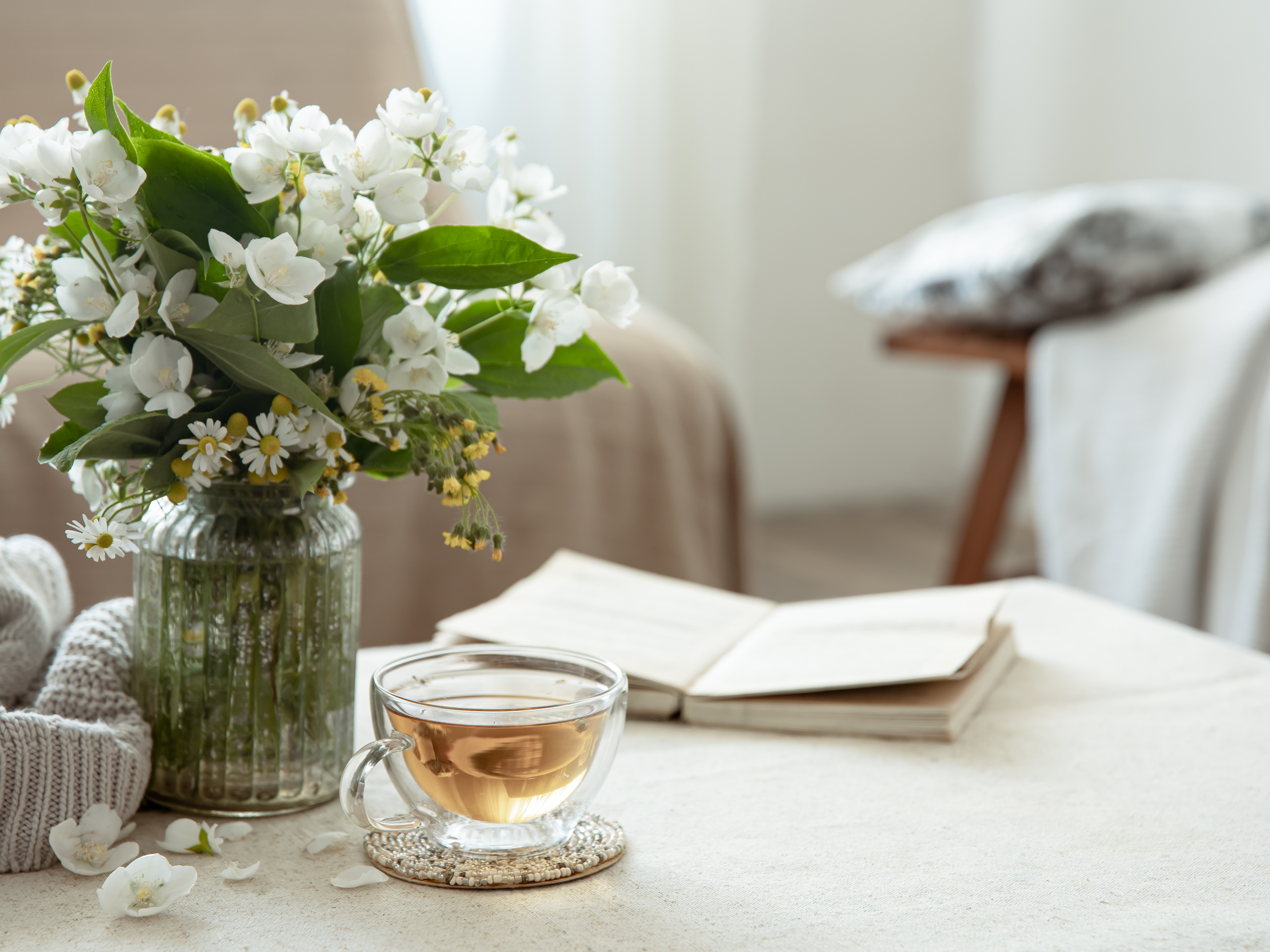 温かいハーブティーが入ったガラスのカップと、白い花を活けたガラスの花瓶が並ぶテーブル。そばには開いた本と柔らかいニットがあり、リラックスした雰囲気が漂う。