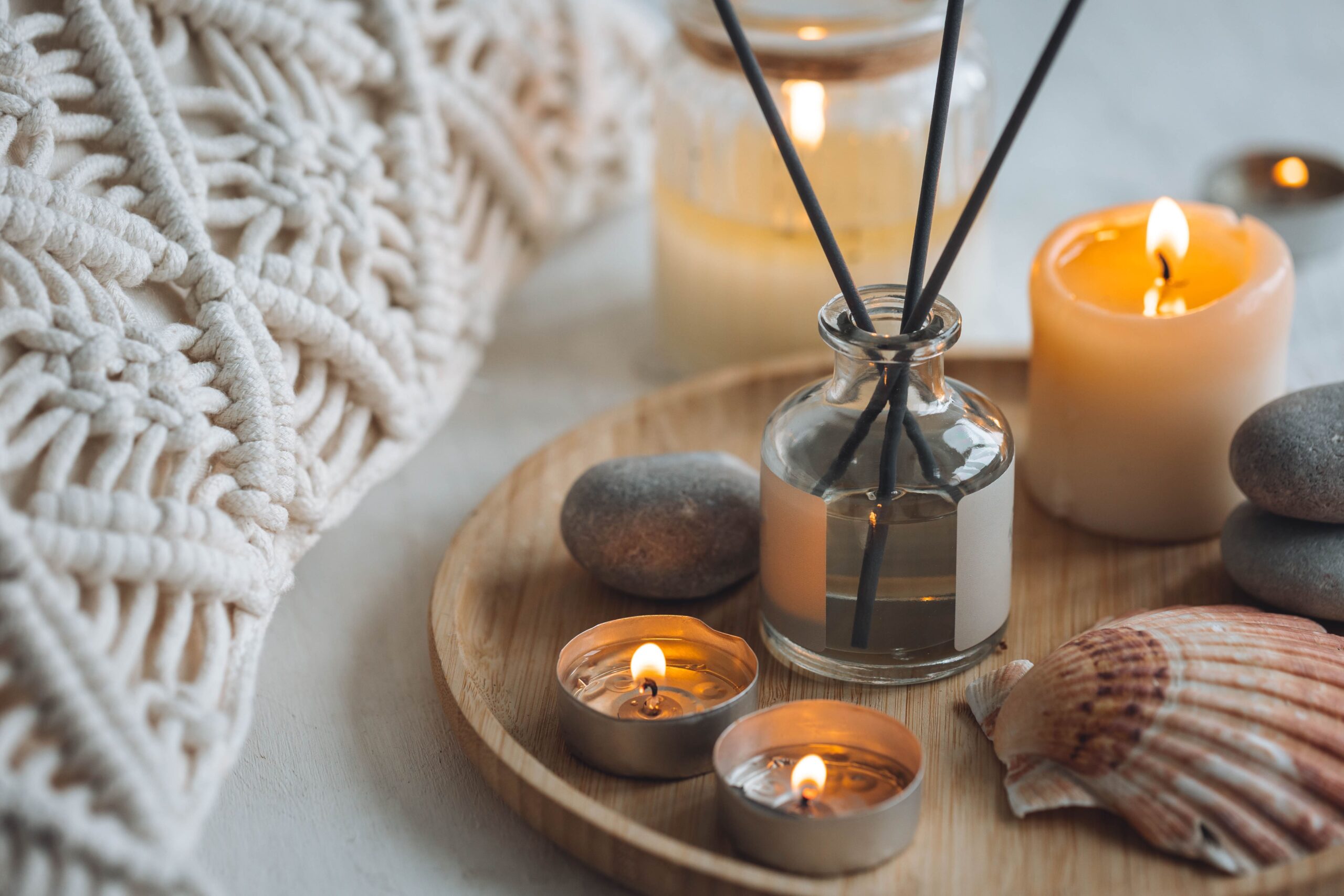 木製のトレイに置かれたアロマディフューザーと、優しく灯る複数のキャンドル。マクラメ編みのクッションが添えられた、ヒュッゲでリラックスした空間の演出。