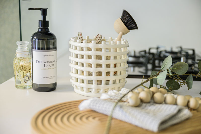 白を基調とした清潔感のあるキッチンカウンター。おしゃれなディッシュソープボトル、白いバスケット、木製のキッチンツールが整然と並び、心が整う空間を表現している。